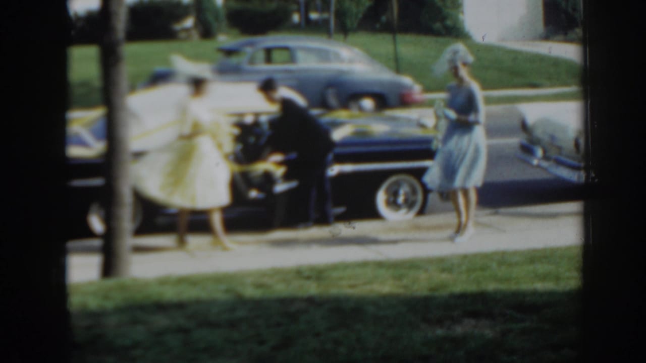 A man helps a woman in a yellow dress beside a decorated black car.