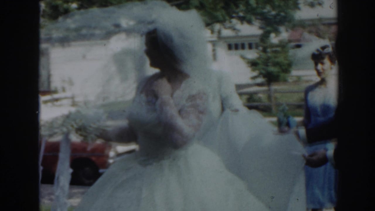 Bride in a white gown and veil approaches a decorated car beside a woman in a blue dress.