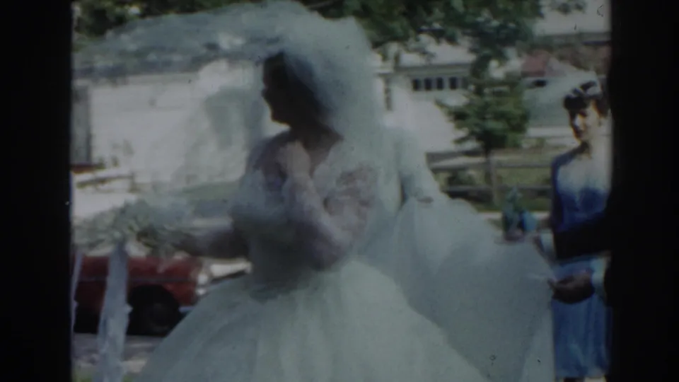 1963 New Jersey 8mm frame showing a bride in a white gown and veil walking toward a ribbon-decorated black sedan with a woman nearby.