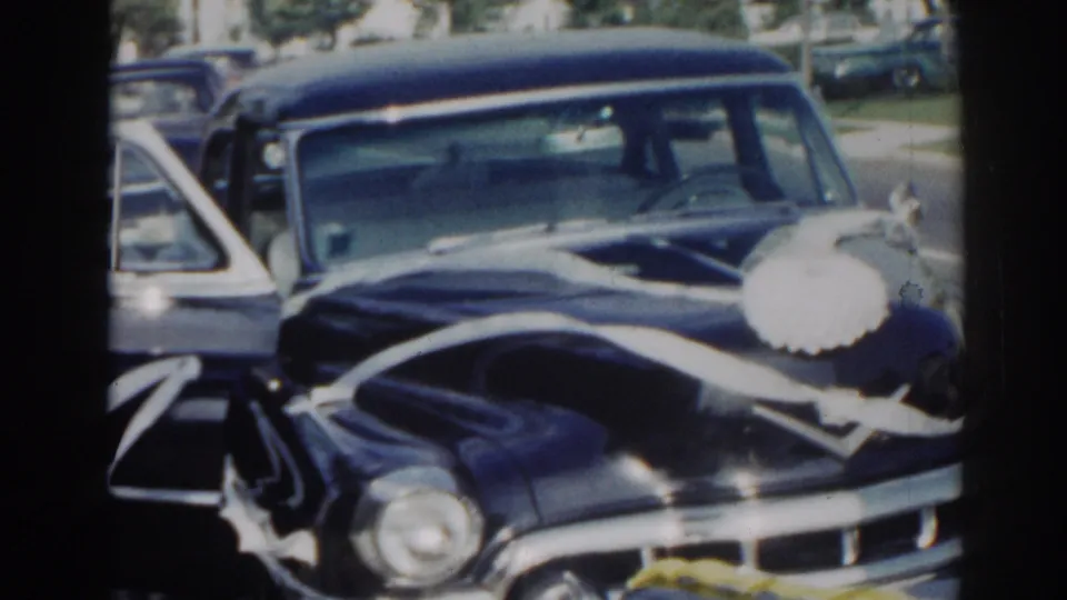 1963 home-movie frame focused on the ribbon-decorated black sedan prepared for a wedding departure in a residential street setting.