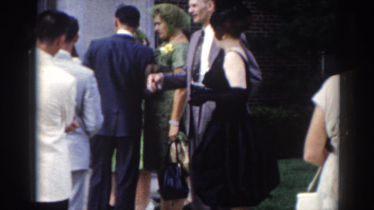 Adults in formal clothing stand outside a light-colored building, with women in dresses and men in suits.