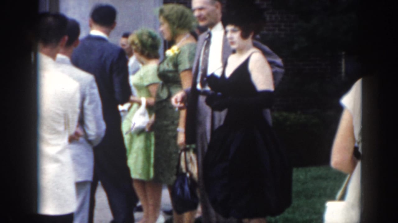 1963 New Jersey frame of adults in suits and dresses gathered outside, including a woman in green and another in a dark dress with gloves.