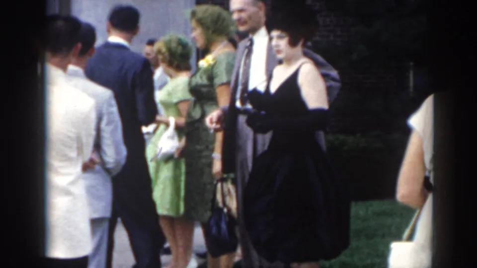 1963 New Jersey frame of adults in suits and dresses gathered outside, including a woman in green and another in a dark dress with gloves.