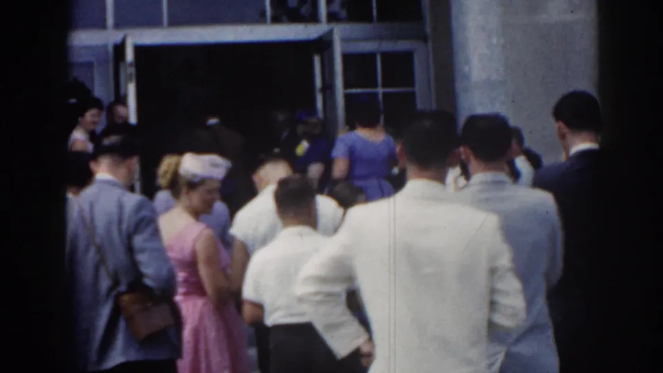 1963 New Jersey home-movie frame showing a small crowd in suits, dresses, and hats gathered near open doors of a light-colored building.
