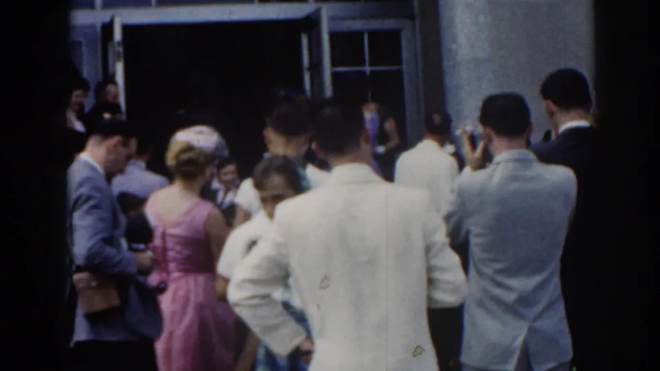 1963 New Jersey frame of formally dressed adults waiting and talking outside an open building entrance, with light jackets and pastel dresses visible.
