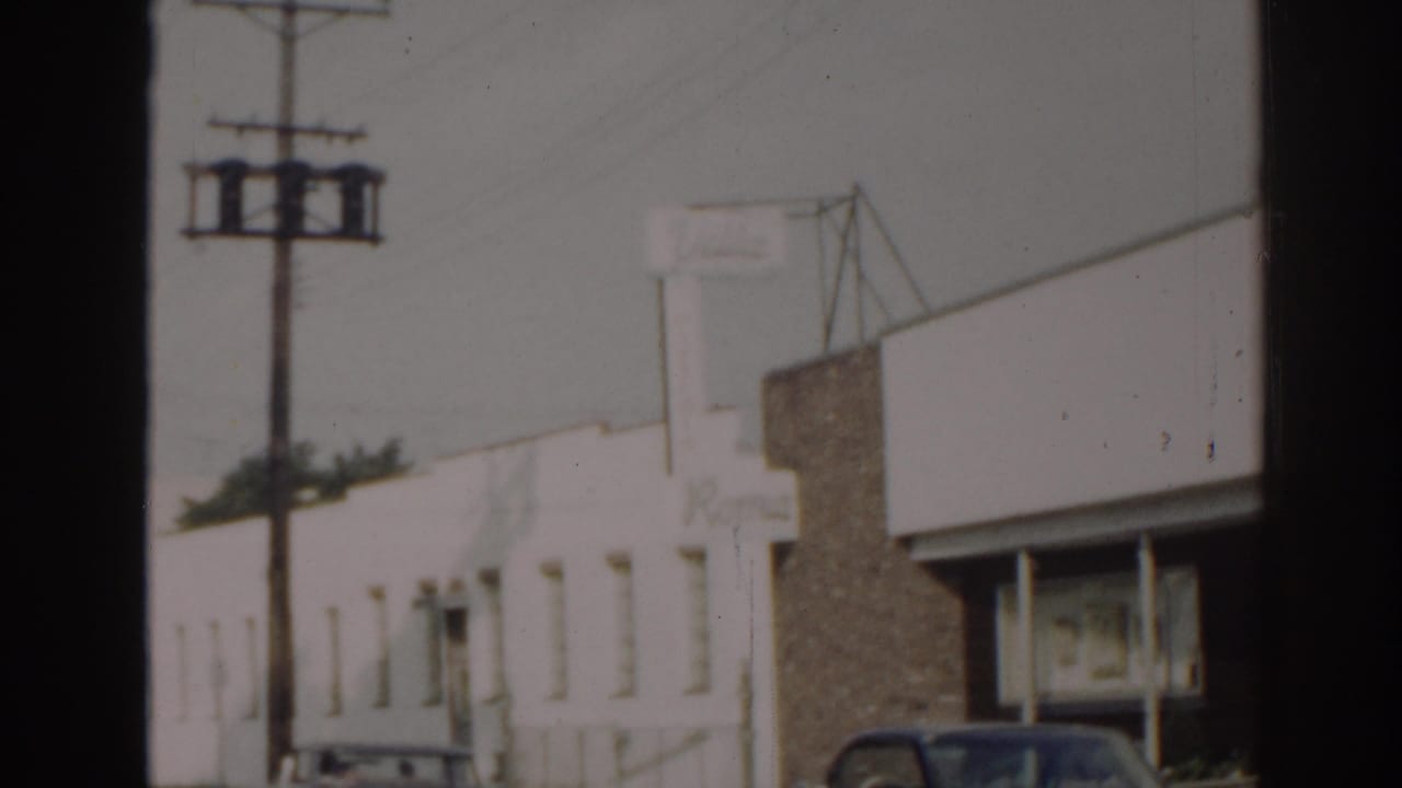 1963 New Jersey 8mm frame showing a white mid-century storefront, brick facade sections, utility pole, overhead wires, and parked cars.