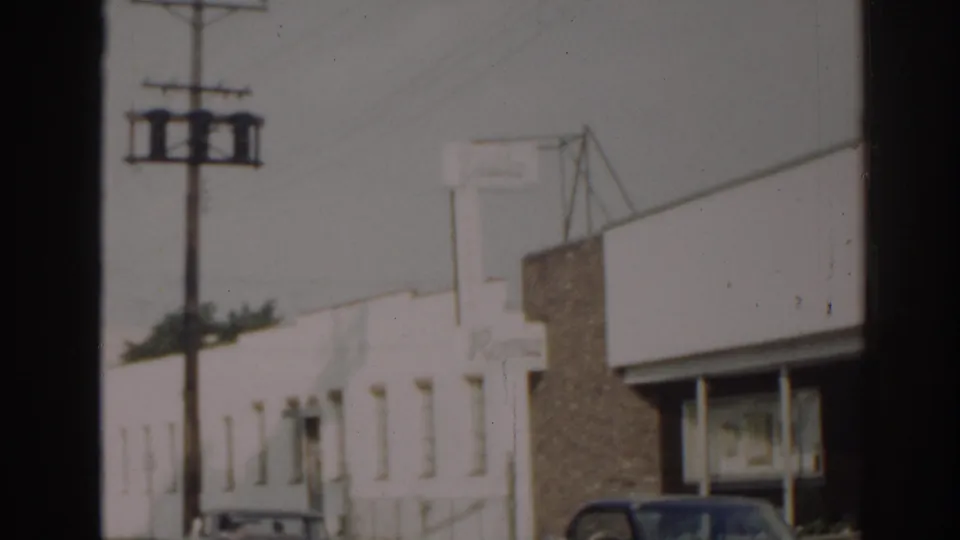 1963 New Jersey 8mm frame showing a white mid-century storefront, brick facade sections, utility pole, overhead wires, and parked cars.