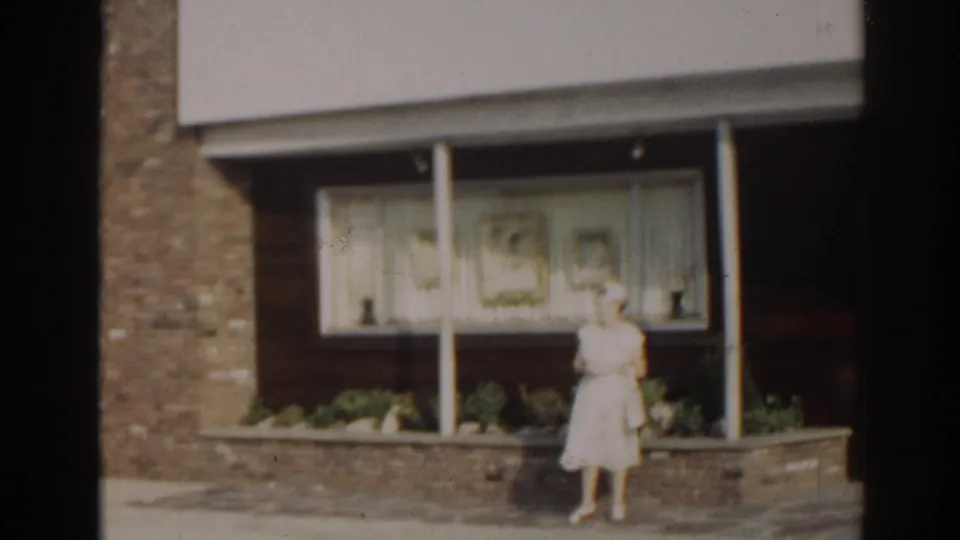 1963 New Jersey frame showing an adult woman in a light sundress standing outside a storefront window with curtains and low planters.