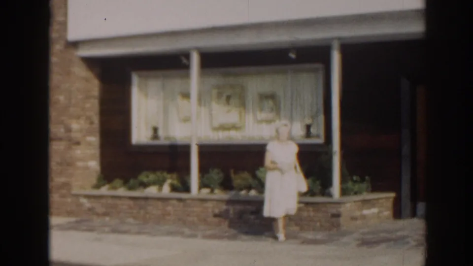 1963 New Jersey 8mm frame of an adult woman outside a mid-century storefront, standing near the entrance beside windows and planters.