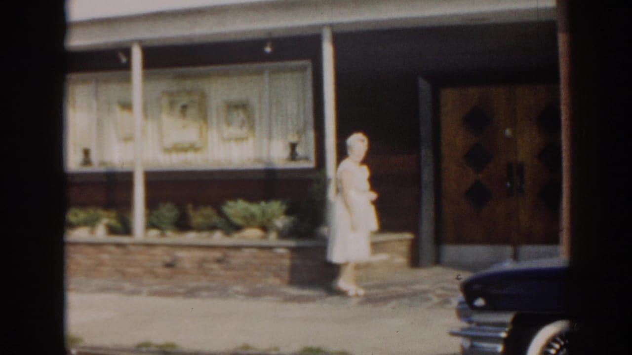 Woman walking past large windows and a wooden doorway with a parked car at the edge of frame.