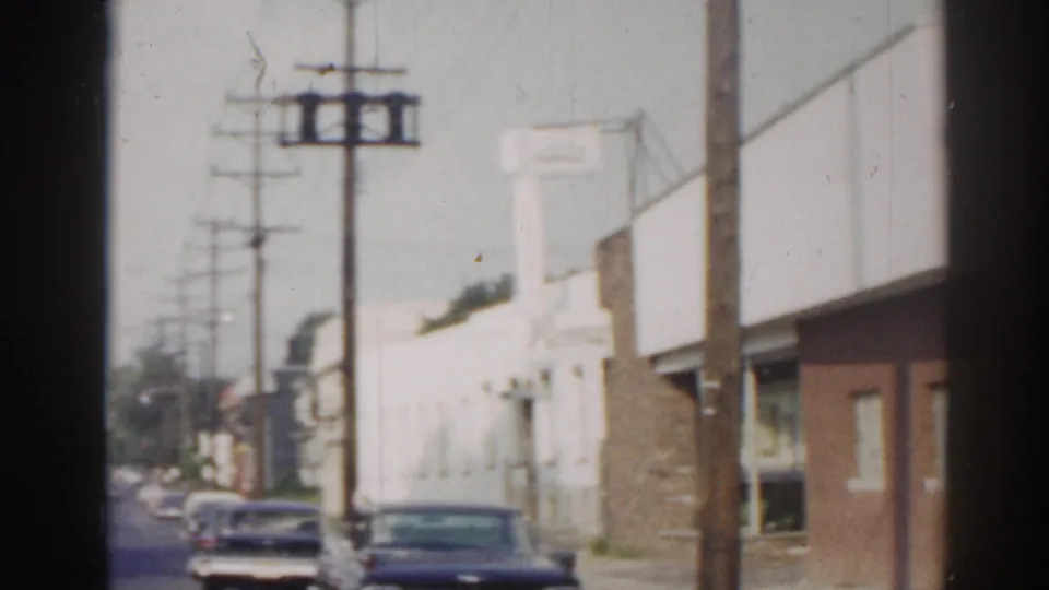 1963 New Jersey home-movie street frame showing utility poles, parked cars, and low commercial or residential buildings.