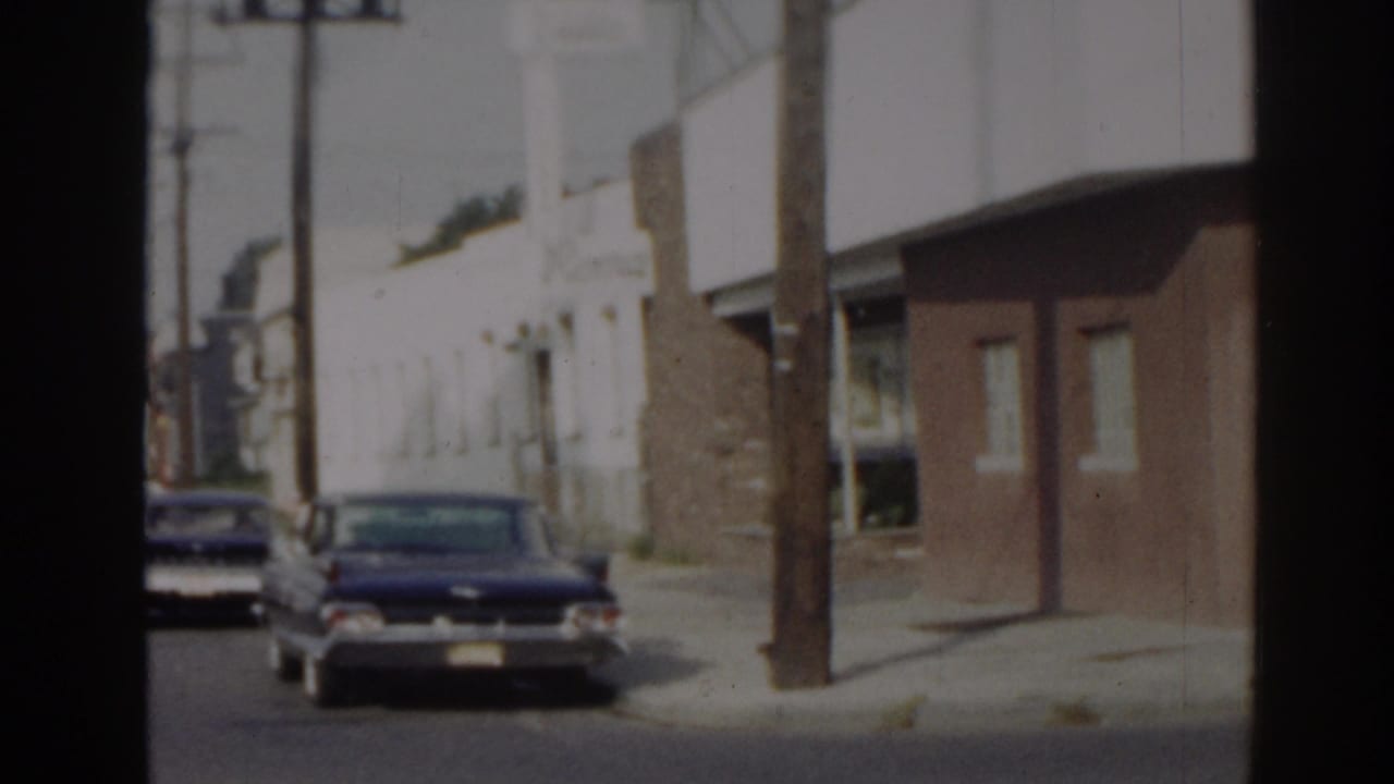 1963 New Jersey 8mm frame showing a parked sedan, utility poles, sidewalk, and low buildings on a quiet street.