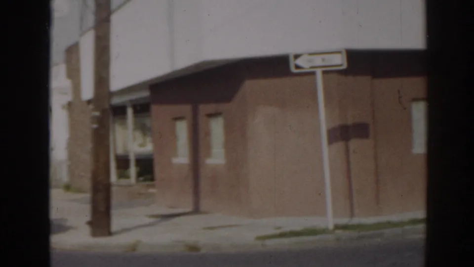 1963 New Jersey 8mm frame showing a brown corner building, sidewalk, pole, and directional street sign.