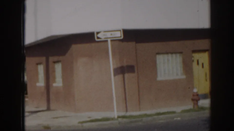 1963 New Jersey street frame with a brown building, yellow door, curtained window, fire hydrant, and directional sign.