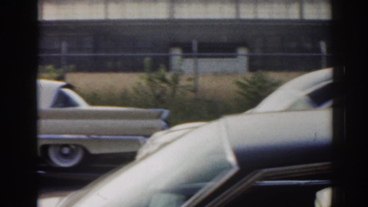 Decorated light-colored cars pass behind a dark window frame near a roadside fence.