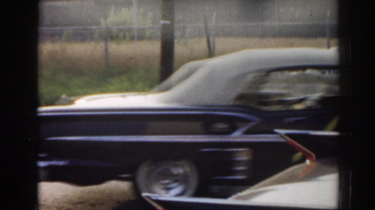 1963 New Jersey 8mm frame with a dark decorated sedan moving past a white car beside roadside grass and fencing.