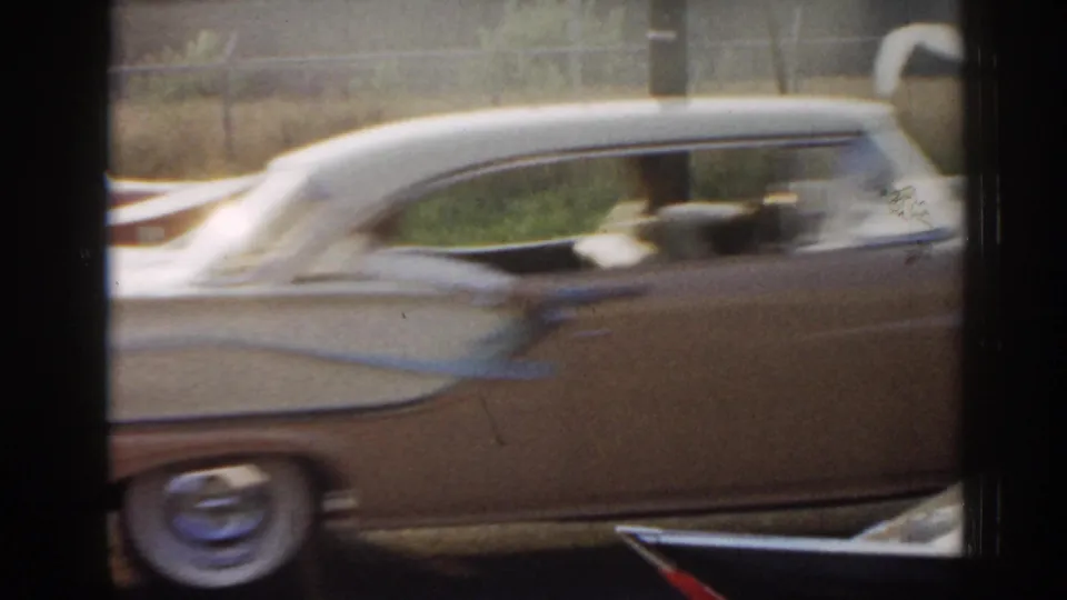 1963 New Jersey 8mm frame showing a light-colored tailfin sedan moving past the camera with a roadside fence beyond.