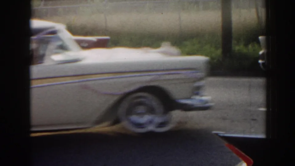 1963 New Jersey 8mm frame with a white decorated car moving past roadside grass and a chain-link fence.