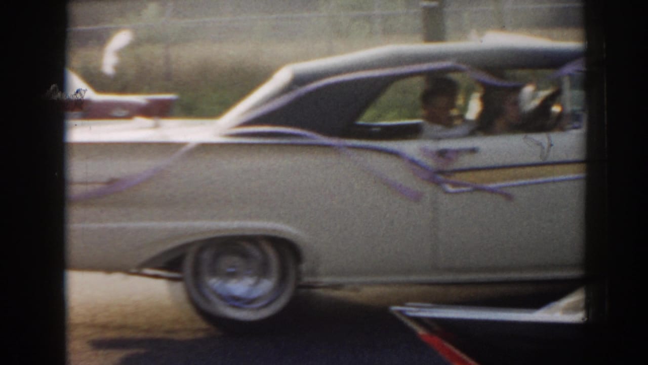 Cream mid-century car with dark roof passing close to camera.