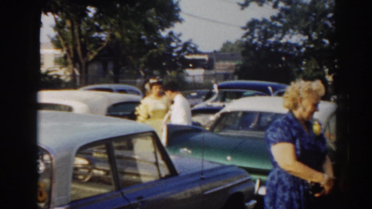 Wedding guests gather in a New Jersey parking lot among early-1960s cars, with a woman in blue at the right edge.