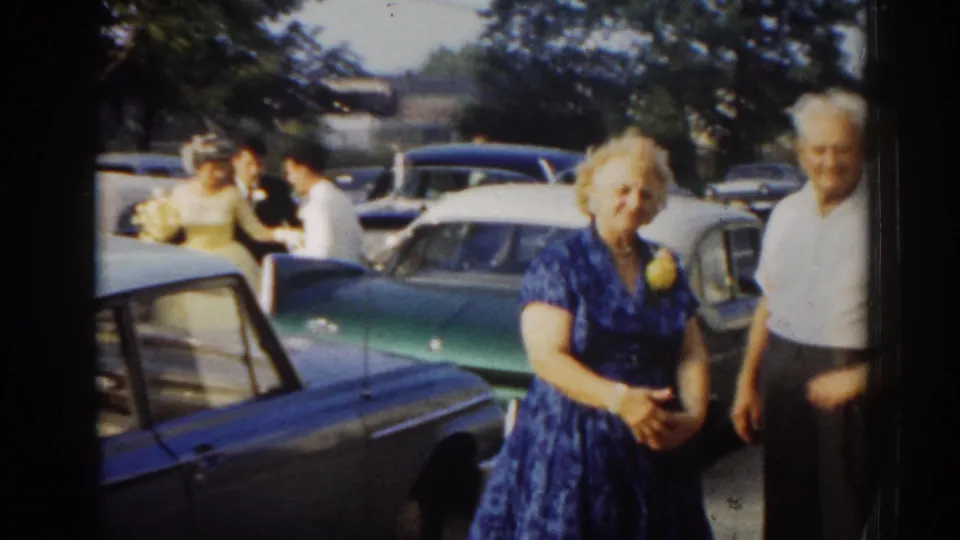 A woman in a blue patterned dress and an older man stand near green and blue classic cars during the 1963 New Jersey wedding gathering.