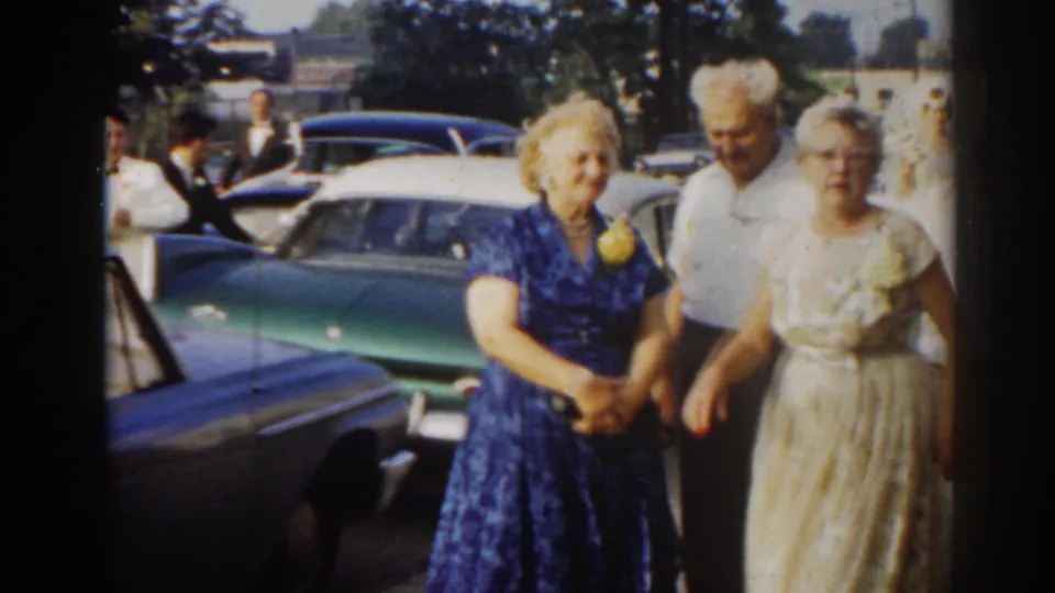 Bride in a white gown stands with two older guests near classic cars in the New Jersey wedding parking lot.