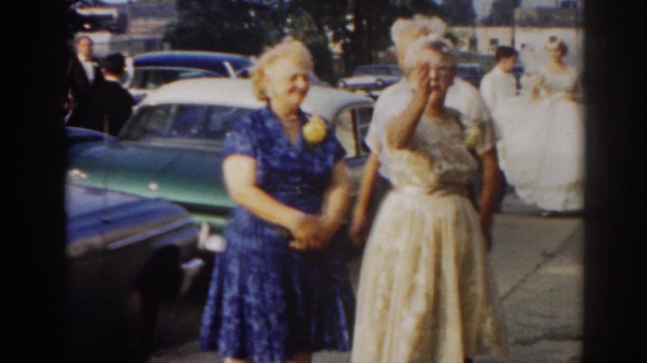 Two older women in formal dresses stand on a sidewalk as a bride approaches in the background.