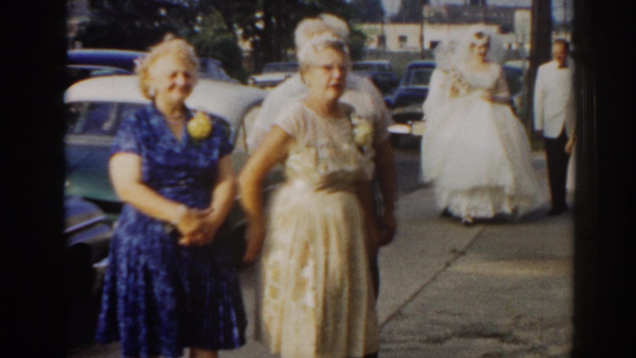 Two women stand in front as the bride in a white gown and other guests approach along the sidewalk.