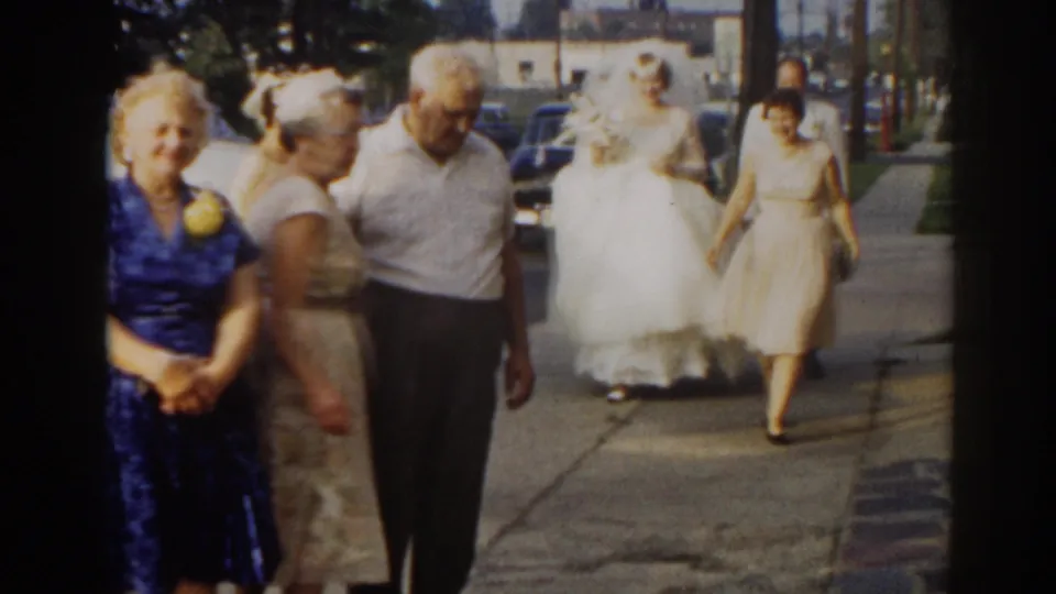 1963 New Jersey home-movie frame showing wedding guests on a sidewalk, with the bride and another woman approaching from the background.