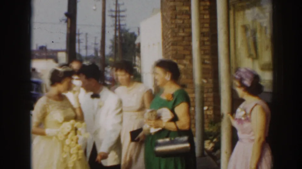 1963 New Jersey wedding frame showing the bride and groom with guests gathered near a brick building entrance.