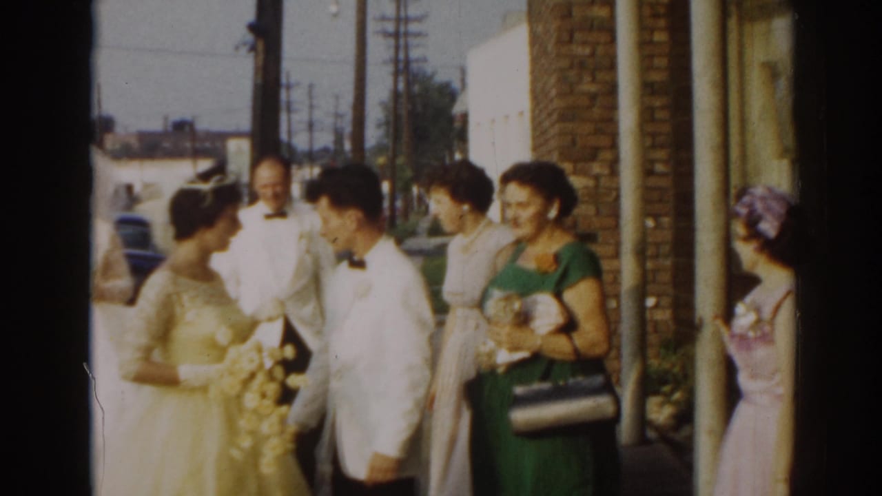 Bride and groom stand with guests outside a brick building on a sunny street.