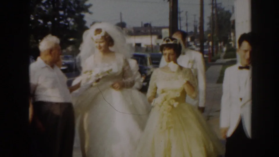 The bride moves along the street-side walkway with an older man, a bridesmaid in pale yellow, and the groom close behind.