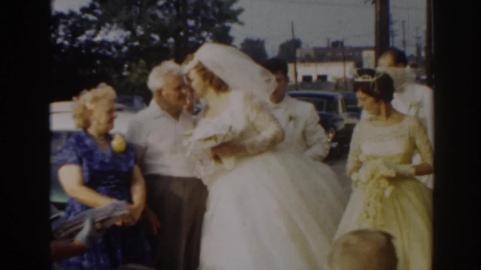 The bride appears to kiss or greet an older man while family members and a bridesmaid stand nearby outside on the street.