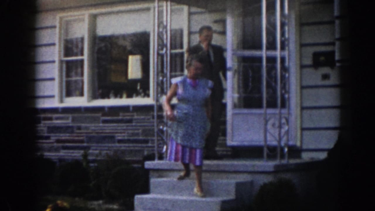 Woman steps down from a suburban front doorway while a suited man stands behind her.