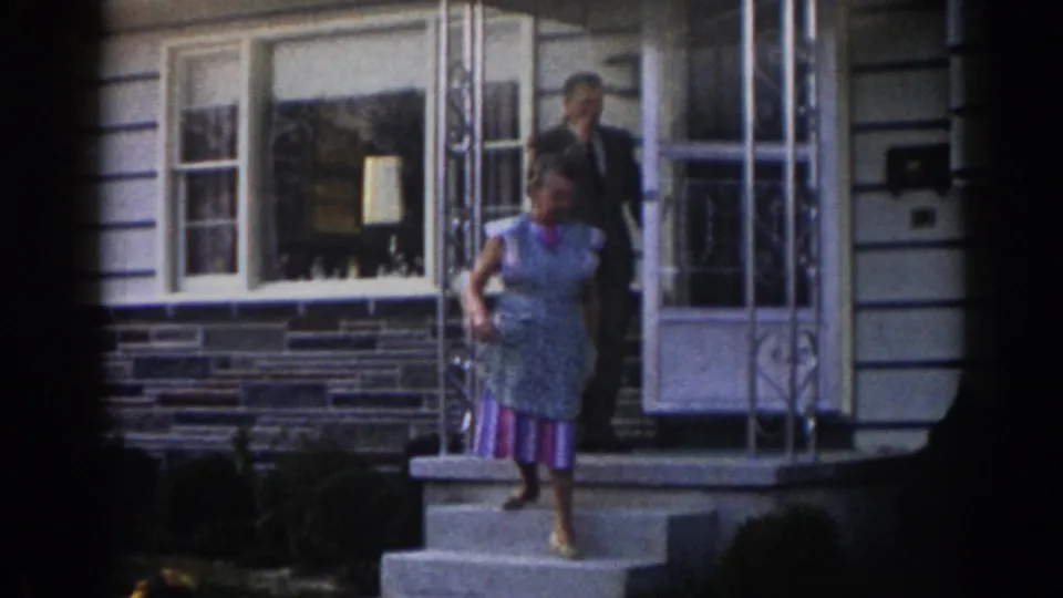 In 1963 New Jersey home-movie footage, a woman in a patterned dress and apron descends the front steps of a suburban house as a man in a suit follows from the doorway.