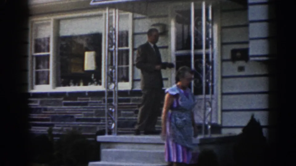 The woman has moved toward the right side of the frame while the suited man remains near the open front door of the New Jersey suburban house.