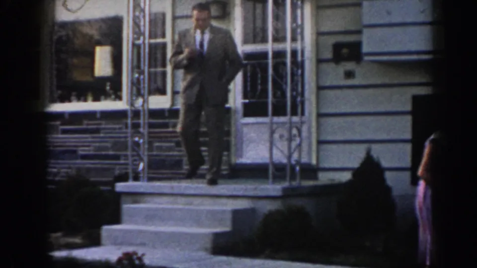 A man in a suit descends the front steps of the suburban house, with the open doorway, window, stone foundation, and partial figure of the woman visible nearby.