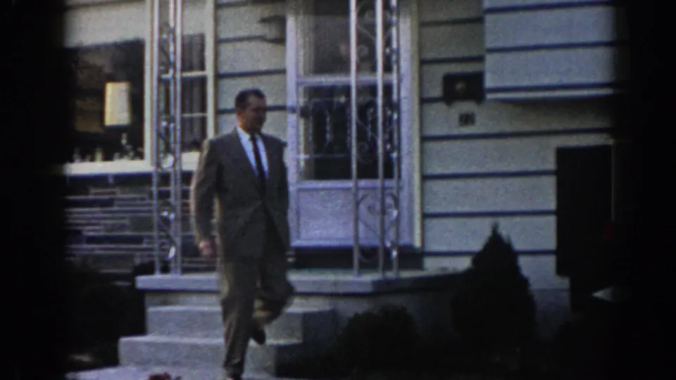 The suited man walks away from the front steps of the New Jersey house, with the open door, porch supports, siding, and dark foreground framing still visible.