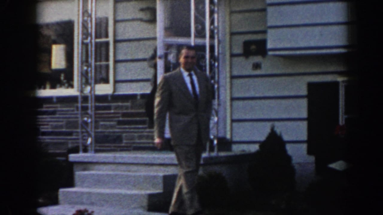 In supported 1963 New Jersey footage, a suited adult man walks away from a residential porch as another figure stands near the doorway.