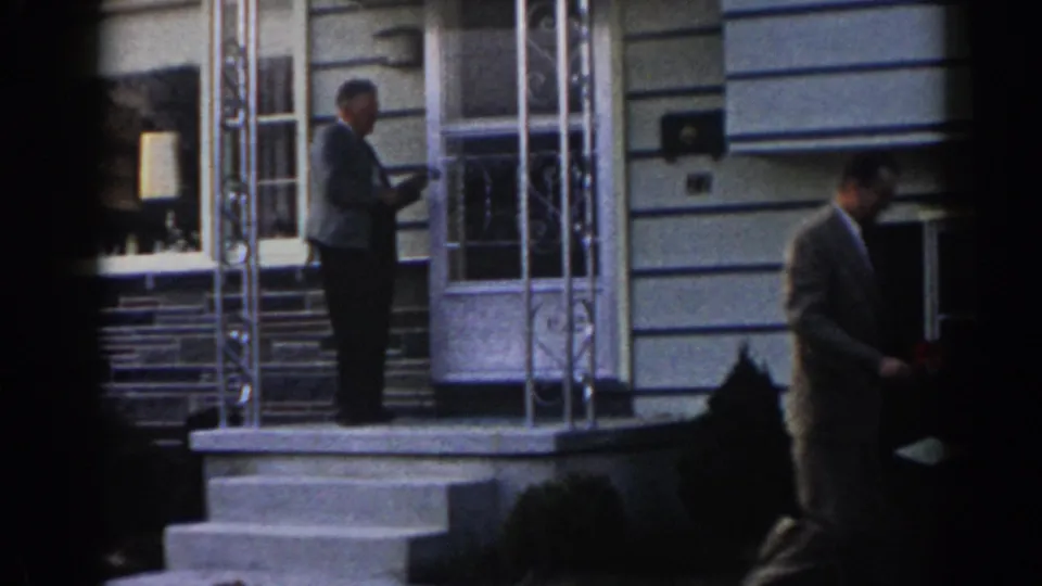 A residential porch scene shows one adult man by the door and another suited man descending or leaving the steps in 1963 New Jersey home-movie footage.