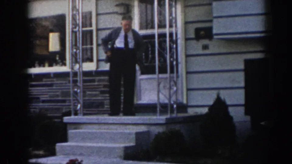 An older adult man stands facing the camera on the front porch of a New Jersey residence, wearing a shirt, tie, and dark trousers.