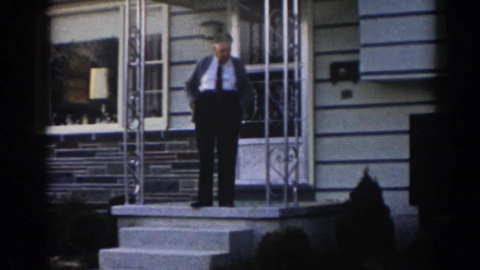 The older man remains on the residential porch in supported 1963 New Jersey footage, framed by the front door, windows, railings, and steps.