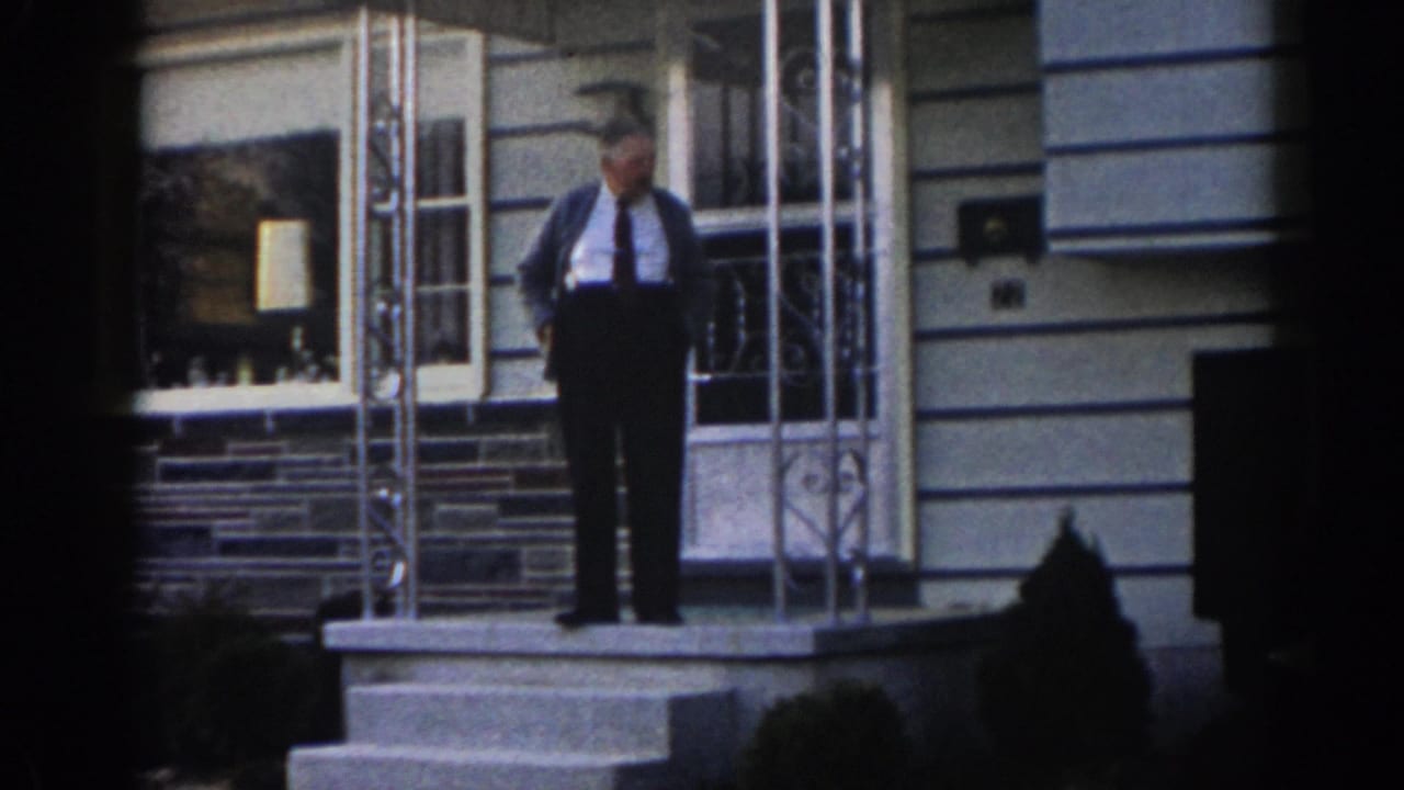 Man in shirt, tie, and cardigan standing on front porch steps.