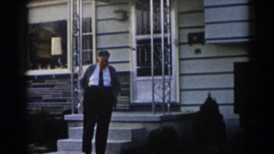 Adult man in shirt and tie shifts his weight on the porch steps outside a 1963 New Jersey suburban house.