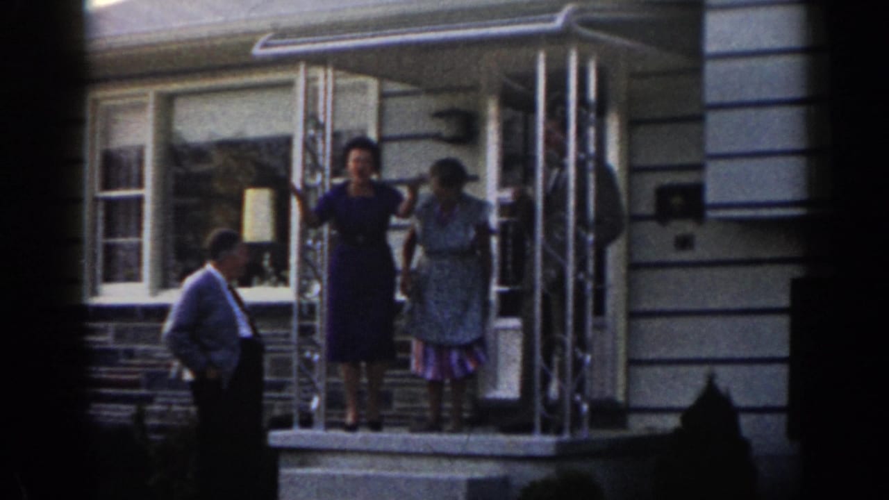 In 1963 New Jersey home-movie footage, two women stand at a residential doorway while a man in a shirt and tie watches from beside the porch steps.