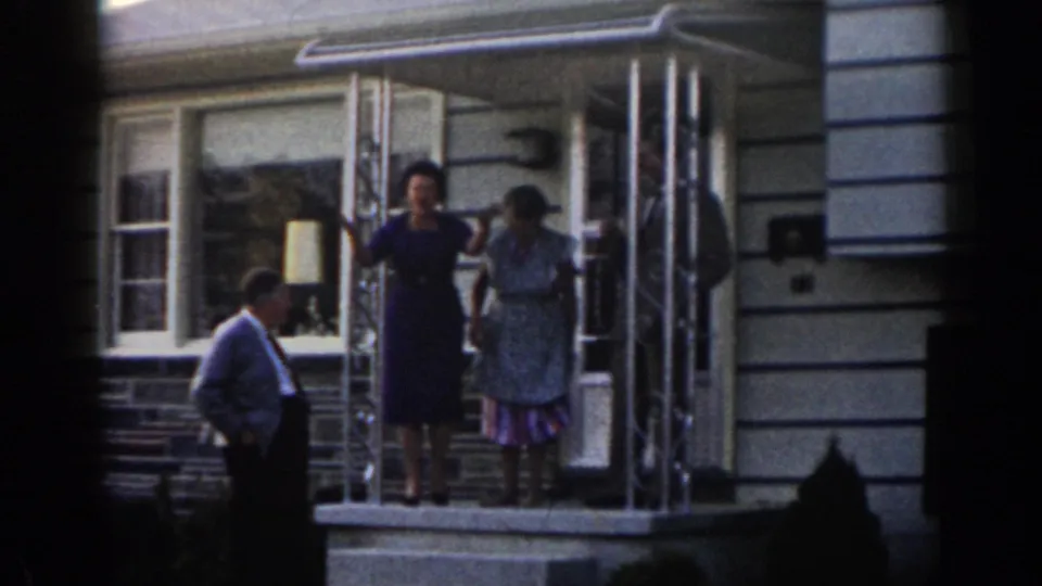 In 1963 New Jersey home-movie footage, two women stand at a residential doorway while a man in a shirt and tie watches from beside the porch steps.