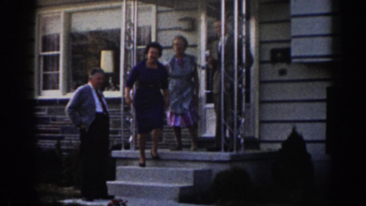 Two women stand on the porch steps while a man stands nearby in front of the house.