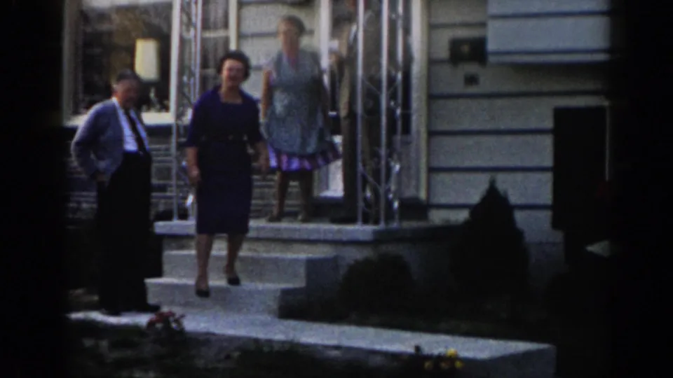 A woman in a dark dress steps from the porch toward the walkway, with another woman still by the door and a man standing beside the steps.