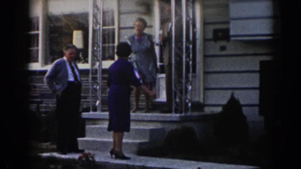 Near the end of the clip, the woman in the dark dress turns back toward the porch while the other woman remains by the doorway and the man stands at the side.