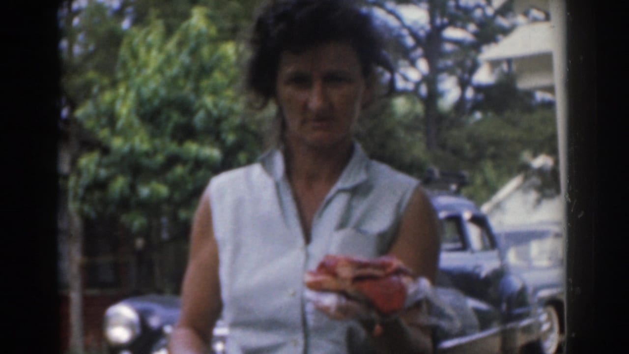 In 1963 New Jersey home-movie footage, a woman holds a wrapped sandwich with trees and a parked vintage car behind her.